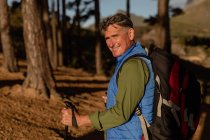 Vista lateral de perto de um homem caucasiano maduro usando uma mochila e segurando bengalas nórdicas de pé e girando para a câmera sorrindo ao sol, durante uma caminhada — Fotografia de Stock