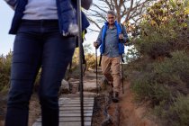 Front view close up of a mature Caucasian man and woman wearing backpacks and using Nordic walking sticks walking single file downhill on a trail during a hike. — Stock Photo