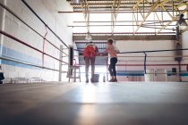Treinador e boxeador feminino interagindo uns com os outros no ringue de boxe no centro de fitness. Forte lutador feminino no treinamento de ginásio de boxe duro . — Fotografia de Stock