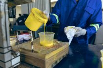 Front view mid section of man working at a cricket ball factory pouring yellow rubber into a plastic container. — Stock Photo