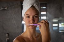 Porträt einer jungen kaukasischen Frau, die sich die Zähne putzt, die Haare in ein Handtuch gehüllt und in einem modernen Badezimmer direkt in die Kamera blickt. — Stockfoto