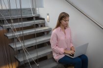 Вид сбоку на молодую белую женщину в розовой рубашке, сидящую на лестнице в квартире с помощью ноутбука . — стоковое фото