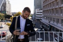 Vue de face d'un jeune homme caucasien souriant tenant un café à emporter et vérifiant l'heure sur sa montre, debout sur une passerelle au-dessus d'une route de la ville. Nomade numérique en mouvement . — Photo de stock