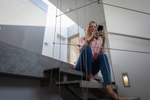 Vista de ángulo bajo de una joven mujer caucásica con una camisa rosa, sentada en una escalera usando un teléfono inteligente . - foto de stock