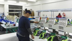 Vista lateral de una joven mujer de raza mixta que opera una máquina de coser automatizada, coser camisas en una fábrica de ropa deportiva, en el fondo son otros colegas que también trabajan con las máquinas . - foto de stock