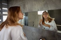 Вид сбоку на молодую белую женщину в халате, расчесывающую волосы, смотрящую в зеркало в современной ванной комнате . — стоковое фото