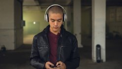Frontansicht eines jungen hispanisch-amerikanischen Mannes, der eine schwarze Lederjacke über einem kastanienbraunen Hemd und Kopfhörer trägt, während er ein Mobiltelefon in einer leeren Lagerhalle benutzt — Stockfoto