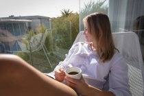 Primer plano de una joven mujer caucásica vestida con una camisa blanca sentada en una silla en un balcón al sol sosteniendo una taza de café y mirando hacia otro lado sonriendo, edificios y árboles en el fondo . - foto de stock