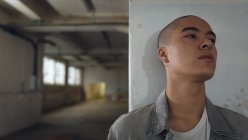 Vista frontal de un joven hispano-americano con piercings con una camisa blanca lisa dentro de una chaqueta gris contra una pared blanca dentro de un almacén vacío mirando lejos de la cámara - foto de stock