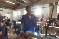 Front view of a young African American man operating a machine in a workshop at a factory making cricket balls looking to camera smiling, in the background a male colleague is sitting at a workbench working on another part of the production line. — Stock Photo