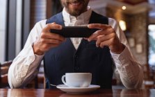 Vue de face au milieu d'un jeune homme caucasien souriant utilisant son smartphone assis à une table avec une tasse de café à l'intérieur d'un café. Nomade numérique en mouvement . — Photo de stock