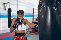 Boxeuse afro-américaine pratiquant la boxe avec sac de boxe dans un club de boxe. Forte combattante dans la boxe gymnase entraînement dur . — Photo de stock