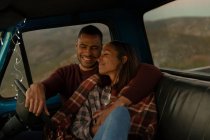 Close up front view of a young mixed race couple sitting in their pick-up truck, smiling and embracing at dusk during a stop off on a road trip. They are sitting in the front seats and the car interior is lit with string lights. — Stock Photo