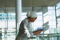 Vista lateral de la mujer de negocios en hijab apoyada en la barandilla y el uso de teléfono móvil en el moderno edificio de oficinas . - foto de stock