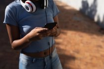 Visão frontal seção média da mulher de pé em um parque urbano usando um smartphone usando fones de ouvido em torno de seu pescoço — Fotografia de Stock