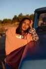 Portrait d'une heureuse jeune femme métissée avec une couverture sur les épaules, appuyée sur le capot d'une camionnette, regardant ailleurs et appréciant l'environnement rural lors d'un arrêt sur la route — Photo de stock