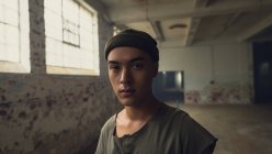 Front view of a young Hispanic-American man with piercings wearing a dark grey shirt and beanie sitting looking intently at the camera while standing inside an empty warehouse — Stock Photo