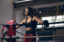 Low angle view of female boxer with eyes closed leaning on ropes in boxing ring at boxing club. Strong female fighter in boxing gym training hard. — Stock Photo