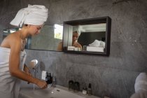 Vista lateral de una joven mujer caucásica con una toalla de baño y con el pelo envuelto en una toalla, sosteniendo un cepillo de dientes, reflejado en un espejo en un baño moderno . - foto de stock