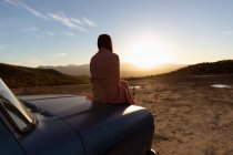 Rückansicht einer jungen Frau, die auf der Motorhaube eines Pick-ups sitzt und die Aussicht bei Sonnenuntergang während eines Stopps auf einer Autoreise genießt — Stockfoto