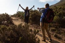Rear view of a mature Caucasian man taking a photo of his partner, a mature Caucasian woman, standing on a trail and raising her Nordic walking sticks in the air during a hike — Stock Photo