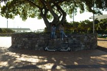 Vue de face de deux jeunes sœurs souriantes de race mixte adultes assis sur un mur dans un parc urbain, avec leurs scooters électriques garés en dessous d'eux — Photo de stock