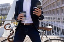 Vorderansicht mittlerer Abschnitt eines Mannes, der einen Kaffee zum Mitnehmen in der Hand hält und ein Smartphone benutzt, auf seinem Fahrrad auf einem Fußweg über eine Stadtstraße gelehnt. Digitaler Nomade unterwegs. — Stockfoto