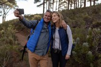 Front view close up of a mature Caucasian woman and man wearing backpacks and a camera, smiling and taking a selfie with a smartphone during a hike — Stock Photo