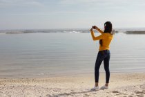 Rückansicht einer jungen Frau mit Kopfhörern, die am Strand steht und mit ihrem Smartphone Fotos vom Meer macht — Stockfoto