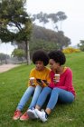 Вид спереду крупним планом дві молодих сестер змішаної раси, які дивляться на свої смартфони і тримають каву, сидячи разом на траві в парку — стокове фото