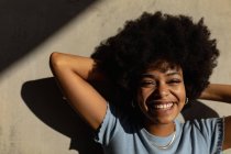 Портрет крупним планом молодої змішаної раси жінки, спираючись назад руками за голову, посміхаючись фотоапарату на відкритому повітрі на сонці — стокове фото