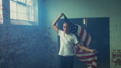 Vista frontal de un joven hispano-americano con una camisa blanca que sostiene una bandera estadounidense dentro de un almacén vacío - foto de stock