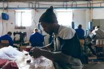 Side view close up of a young African American man wearing a hat sitting at a workbench holding thread in his mouth and working with cut out red leather shapes at a factory making cricket balls — Stock Photo
