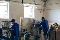 Vista trasera de dos hombres afroamericanos que trabajan en una fábrica haciendo pelotas de cricket, uno está alimentando una pieza de cuero en una máquina . - foto de stock