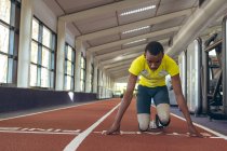 Vista frontal del atlético masculino afroamericano discapacitado en el punto de partida en la pista de atletismo en el gimnasio - foto de stock
