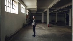 Vue latérale d'un jeune hispano-américain vêtu d'une veste grise, la main dans la poche, regardant la fenêtre, debout dans un entrepôt sombre et vide — Photo de stock
