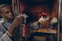 Side view close up of a young African American man standing at a painting area holding a shaped outer half of a red leather cricket ball and spraying it with a spray gun in a workshop at a factory making cricket balls. — Stock Photo