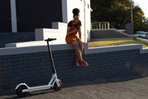 Vista frontale di una giovane donna di razza mista sorridente seduta su un muro per strada con uno smartphone, con il suo scooter elettrico parcheggiato accanto a lei — Foto stock