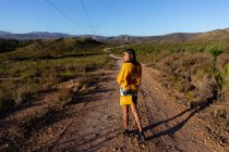Vista laterale di una giovane donna di razza mista che cammina lungo un sentiero attraverso un paesaggio rurale soleggiato e si guarda intorno . — Foto stock