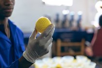 Visão lateral seção média de um jovem afro-americano trabalhando em uma fábrica de bolas de críquete segurando uma bola de borracha amarela de um molde . — Fotografia de Stock
