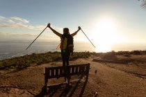 Rear view of a mature Caucasian woman wearing a backpack standing on a bench, raising her Nordic walking sticks in the air and enjoying the view of the coast at sunset during a hike. — Stock Photo