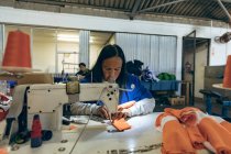 Вид спереди на женщину средних лет, сидящую и работающую на швейной машинке на фабрике спортивной одежды, с коллегой, работающим на швейной машинке на заднем плане . — стоковое фото