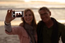 Вид спереду крупним планом зрілого Кавказького чоловіка і жінки посміхаються і приймаючи селфі з смартфоном на пляжі в передній частині моря на заході сонця — стокове фото