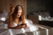 Vista frontal de la joven mujer rubia caucásica acostada en la cama usando un teléfono inteligente . - foto de stock