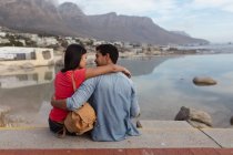 Vista trasera de cerca de una joven pareja de raza mixta sentada fuera en una pared junto al mar abrazándose y mirándose, el mar y las montañas en el fondo - foto de stock
