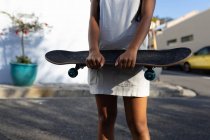 Vista frontale sezione centrale della donna che indossa un vestito, in piedi in una strada urbana che tiene uno skateboard — Foto stock
