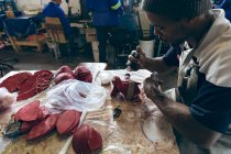 Vista lateral de perto de um jovem afro-americano usando um chapéu sentado em uma bancada de trabalho trabalhando com formas de couro vermelho cortadas em uma oficina em uma fábrica fazendo bolas de críquete — Fotografia de Stock