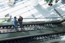 Vista de ángulo alto de hombres de negocios que interactúan entre sí mientras se mueven abajo en escaleras mecánicas en la oficina
. - foto de stock