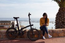 Вид спереду молода змішана жінка, що сидить на вулиці на стіні, використовуючи смартфон з її велосипедом, що спирається поруч з нею, пальма і вид на море на задньому плані — стокове фото