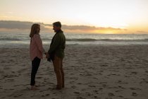 Vista lateral de un hombre y una mujer caucásicos maduros tomados de la mano y parados sonriendo el uno al otro en una playa al lado del mar al atardecer - foto de stock